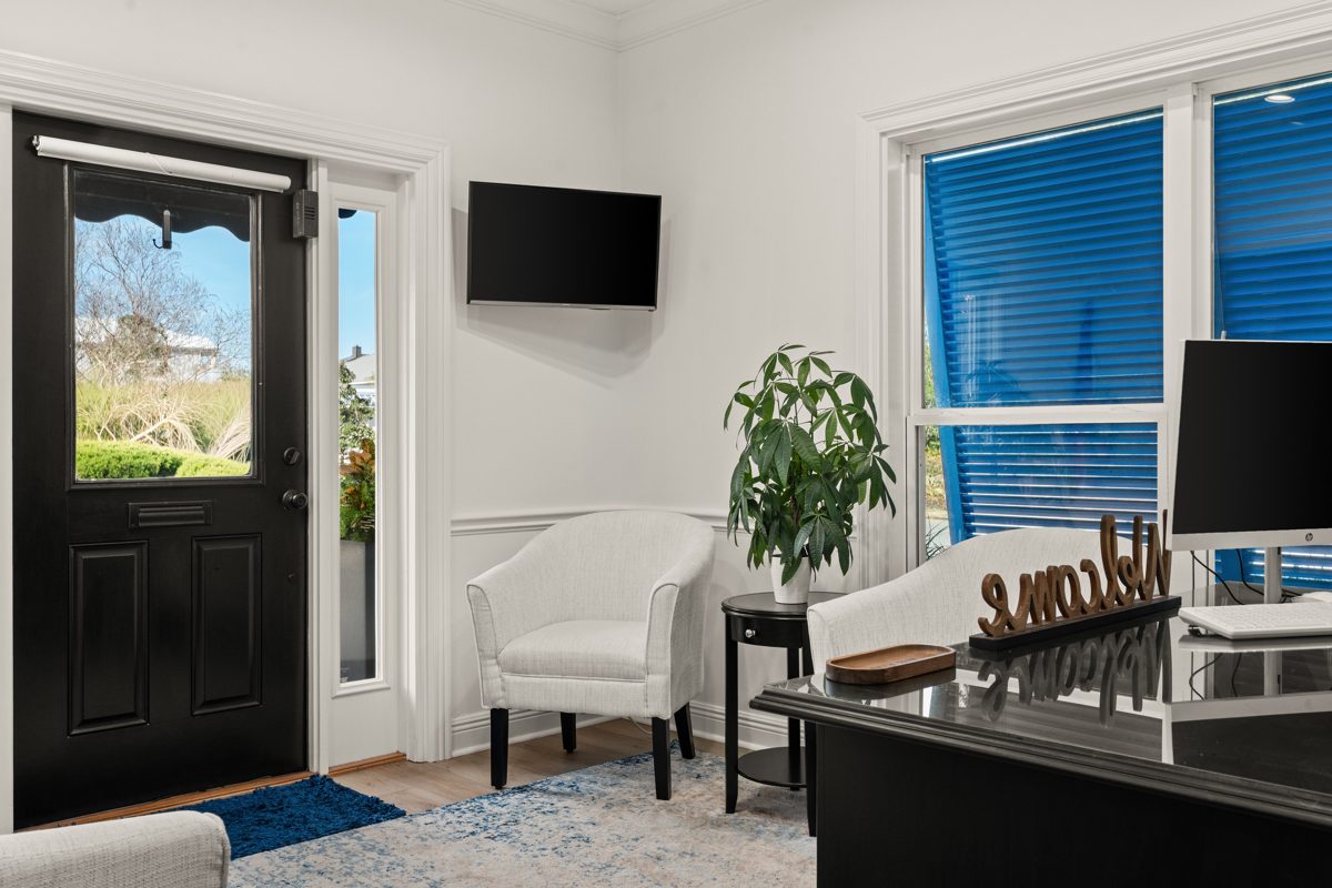 Reception area with a black front door, a white tub chair beside a money tree, and blue shutters glowing through the windows — photo by Jimmy Warr