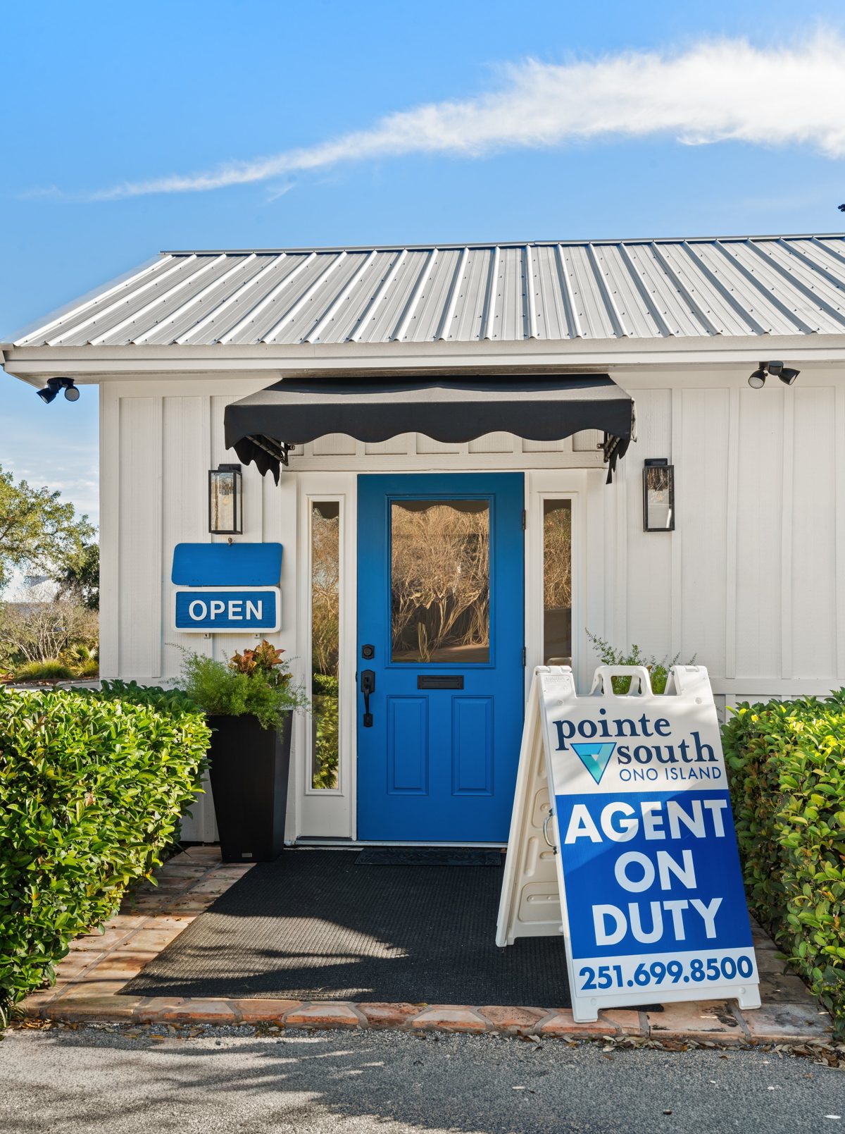 The Pointe South Rentals & Real Estate office on Ono Island: white board-and-batten cottage, blue front door under a black scalloped awning, and an Agent On Duty sign — photo by Jimmy Warr