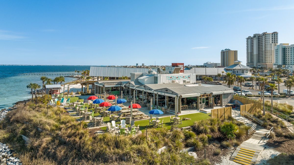 Aerial of the Red Fish Blue Fish patio on Pensacola Beach: red and blue umbrellas over the lawn, dune grass, and Santa Rosa Sound beyond — photo by Jimmy Warr