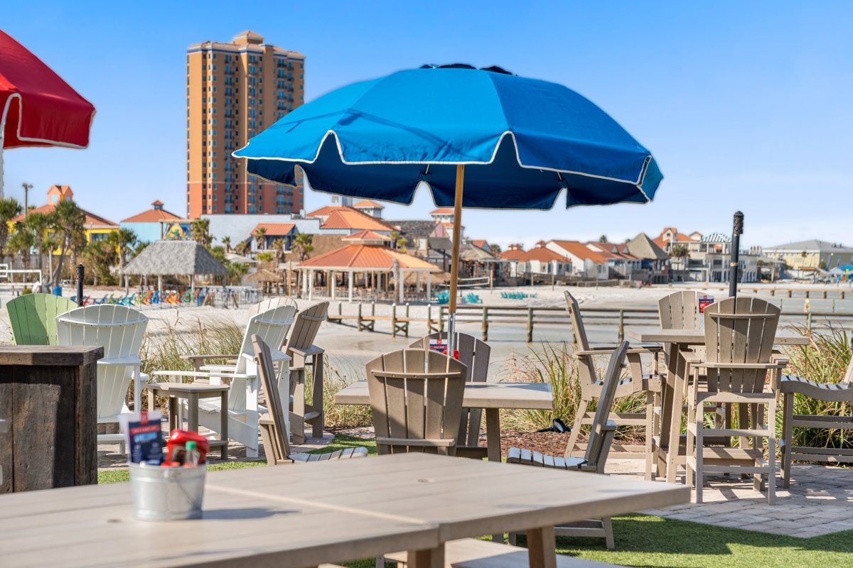 A blue umbrella over patio tables with the beach and boardwalk behind — photo by Jimmy Warr
