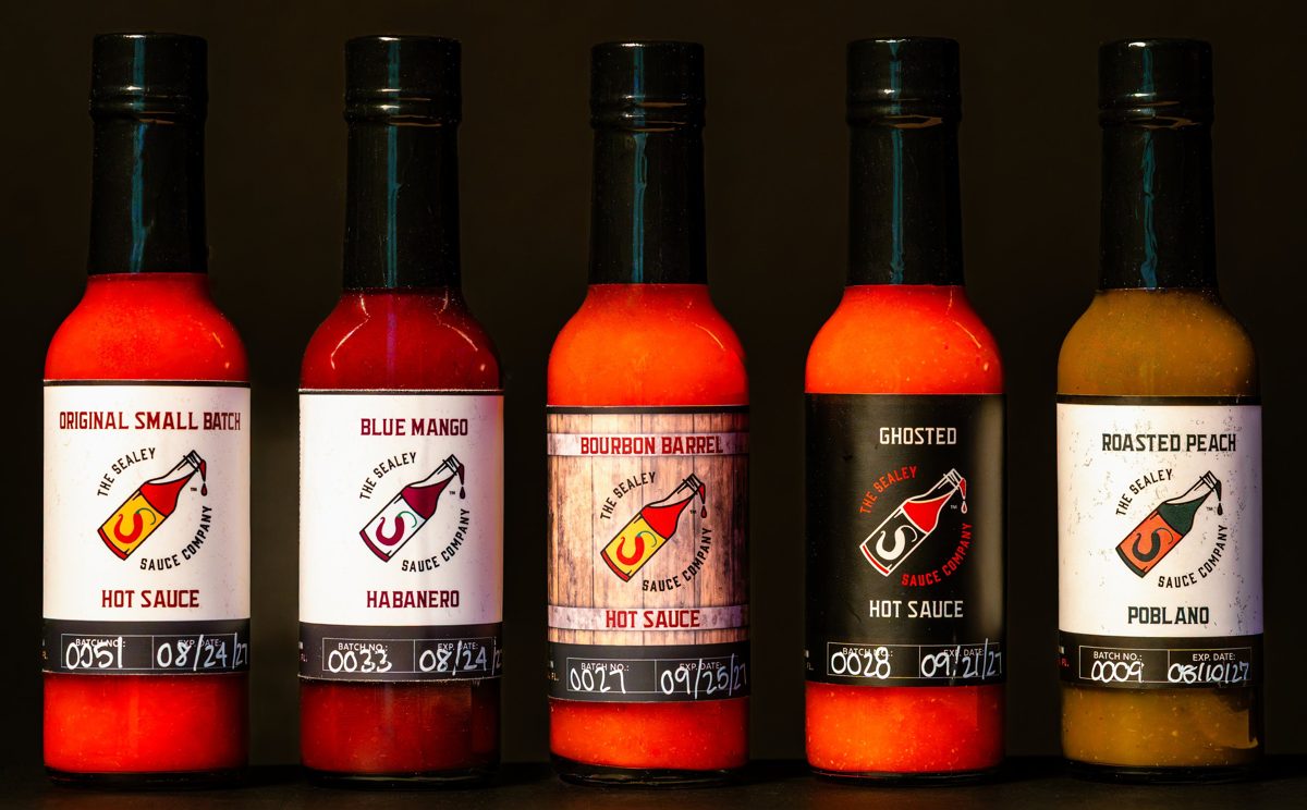 Five The Sealey Sauce Company hot sauces side by side, the Roasted Peach Poblano glowing green-gold at the end of the row — photo by Jimmy Warr