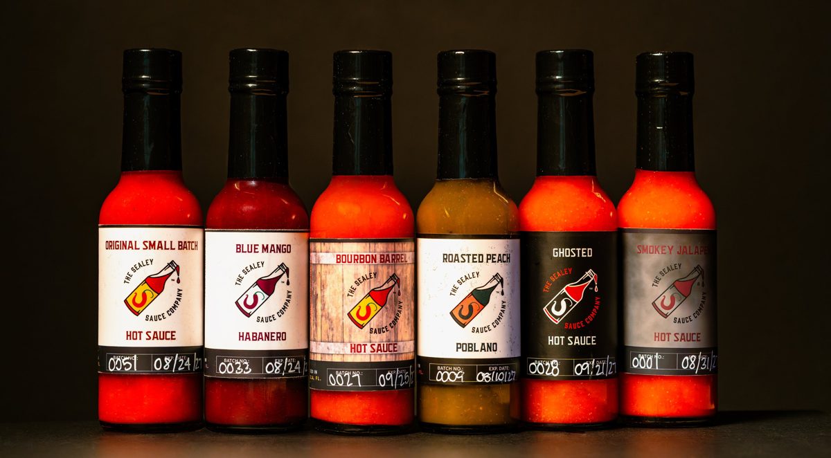Six The Sealey Sauce Company hot sauces lined up on black: Original Small Batch, Blue Mango Habanero, Bourbon Barrel, Roasted Peach Poblano, Ghosted, and Smokey Jalapeño — photo by Jimmy Warr
