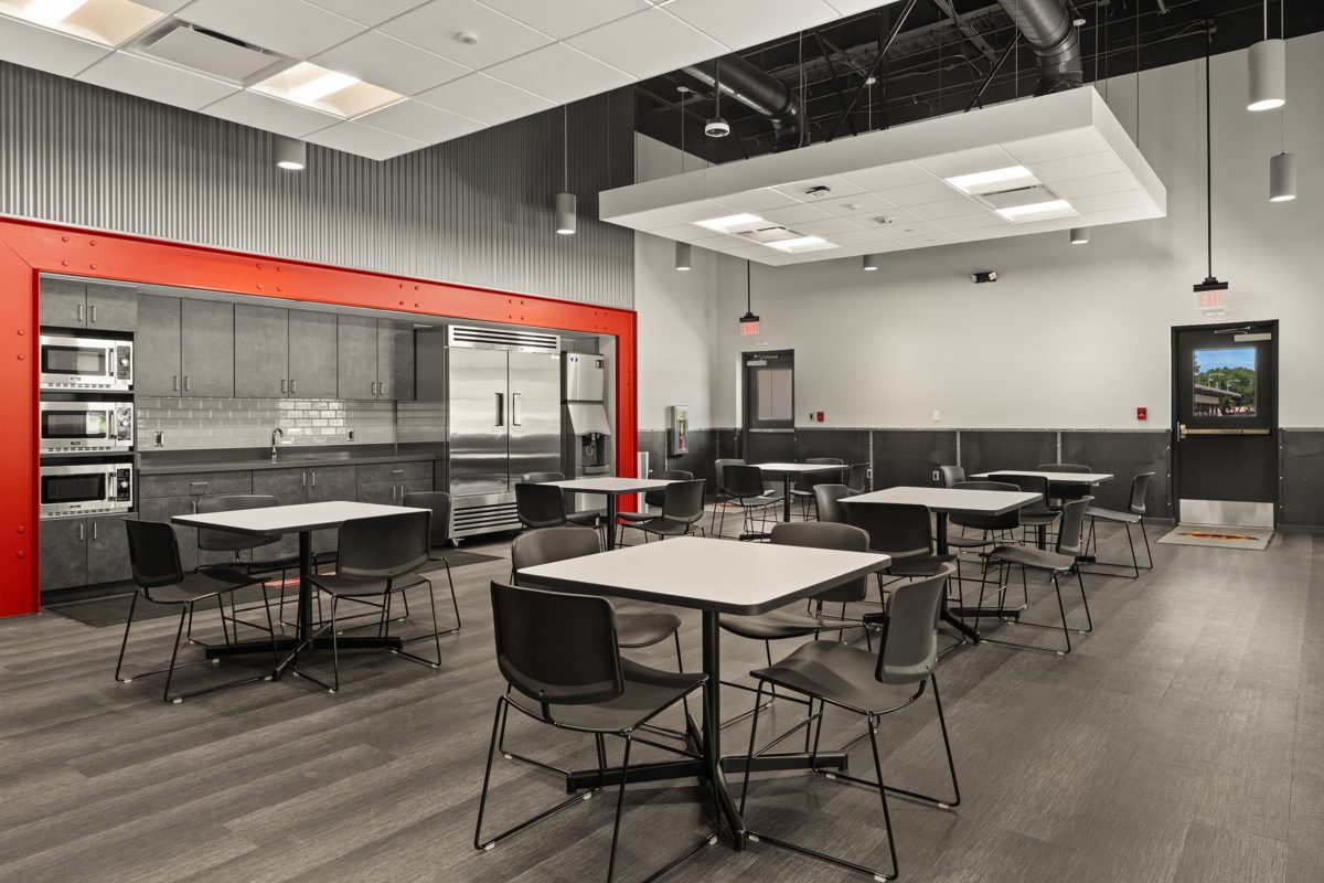 Break room with a red steel-framed kitchen and rows of white tables — photo by Jimmy Warr