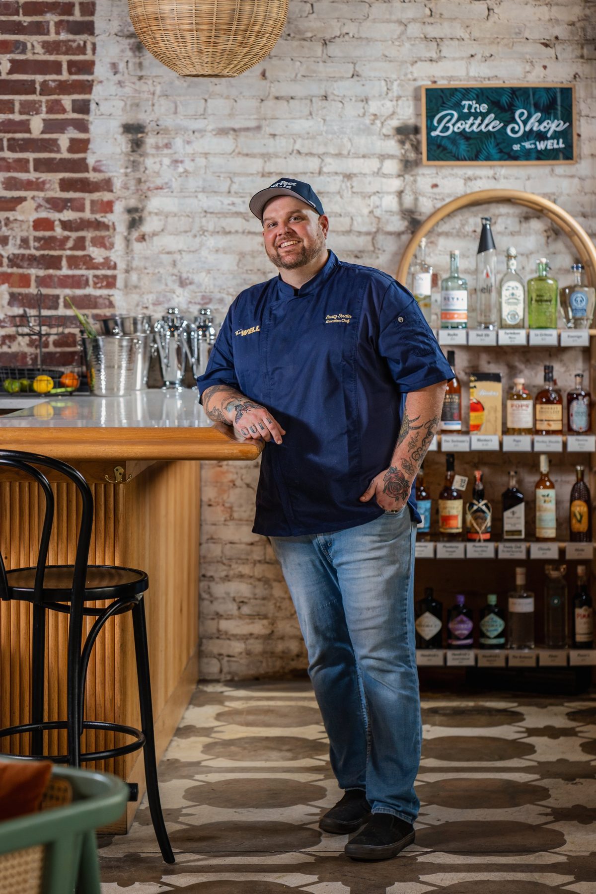 Executive Chef Rusty Strain leans on the bar beside The Bottle Shop at The Well, exposed brick behind him — photo by Jimmy Warr
