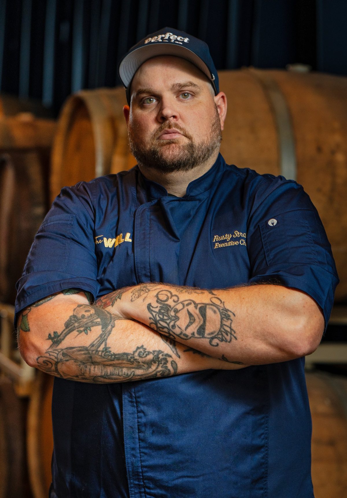 Executive Chef Rusty Strain stands arms crossed in front of wooden barrels — photo by Jimmy Warr