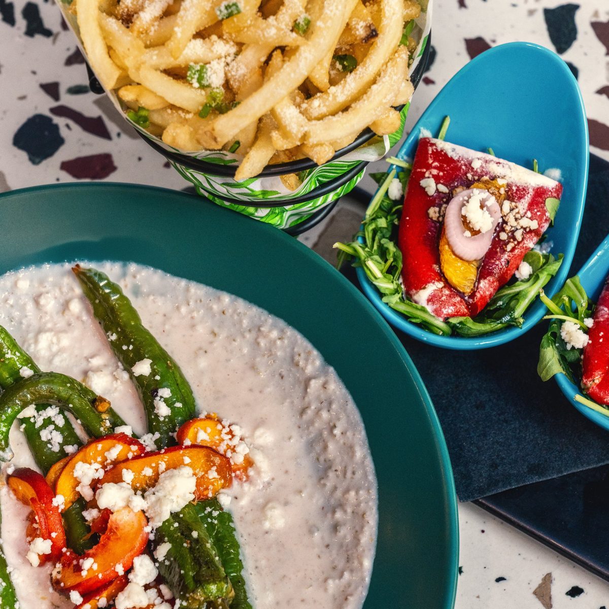 Overhead of dishes from the new menu at The Well: blistered peppers over a creamy base, parmesan fries, and stuffed red peppers on arugula, on a terrazzo table — photo by Jimmy Warr