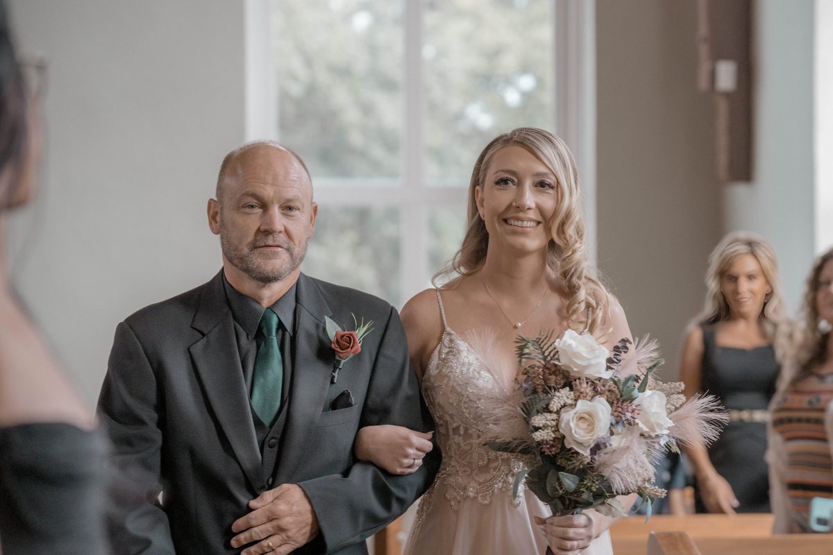 The bride smiles as her father walks her down the aisle, her bouquet of cream roses and pampas in hand — photo by Jimmy Warr