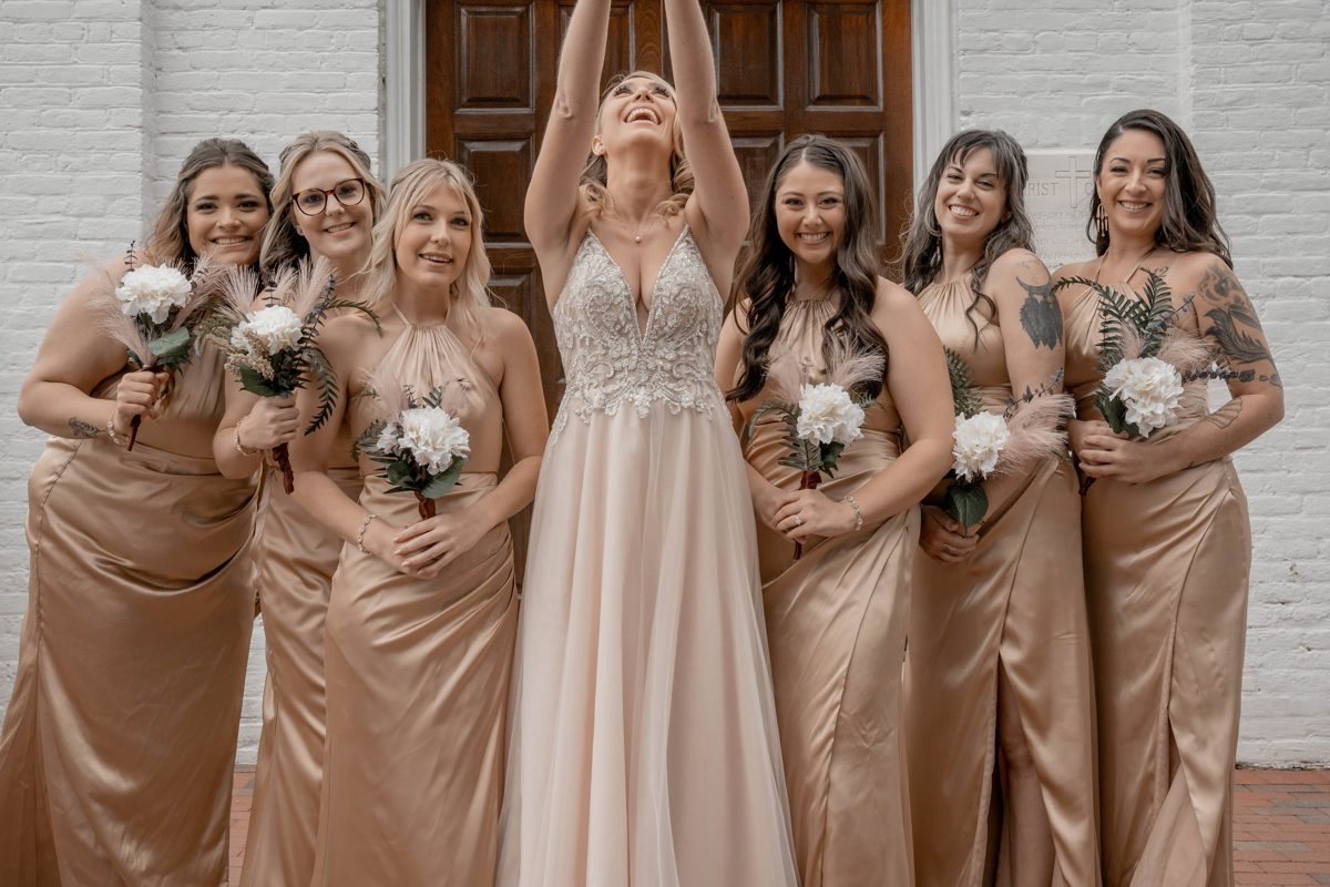 The bride throws her arms up, laughing, surrounded by six bridesmaids in champagne satin — photo by Jimmy Warr