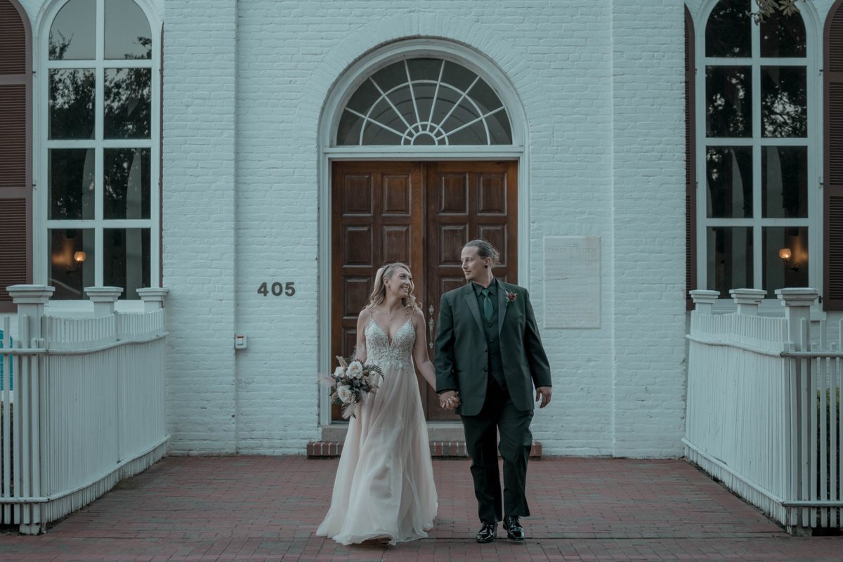 The bride and groom walk hand in hand out of the white-brick front of Old Christ Church in Pensacola, wooden doors and fanlight behind them — photo by Jimmy Warr