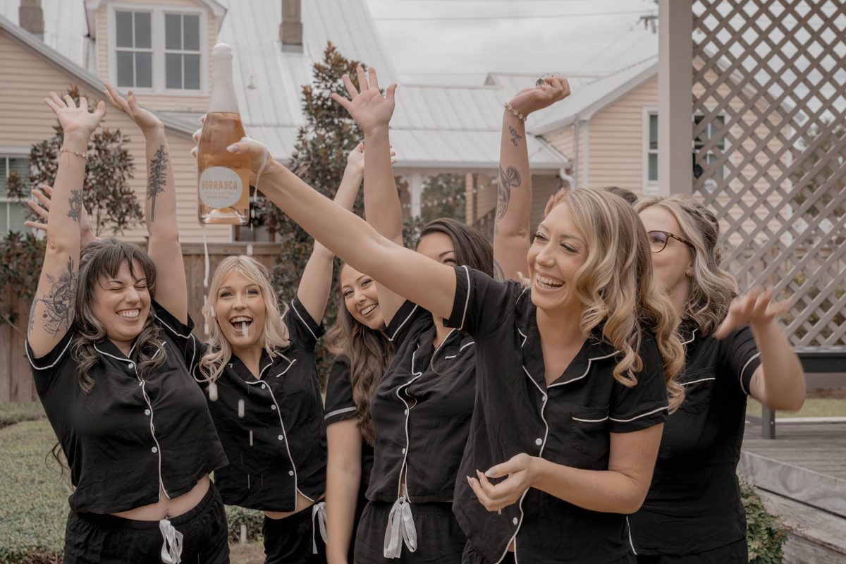 The bride and her bridesmaids in matching black pajamas cheer as a bottle of rosé sprays overhead — photo by Jimmy Warr