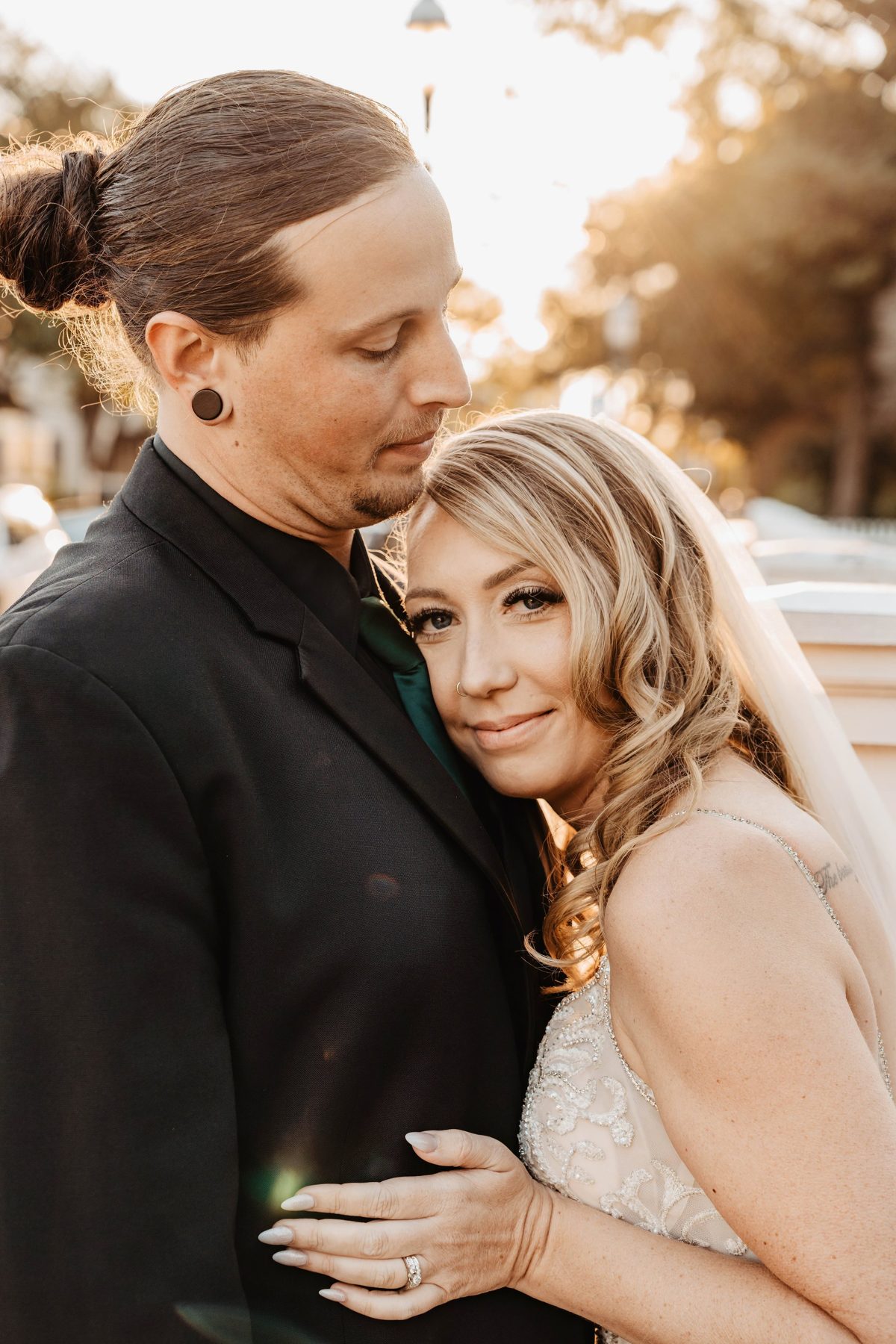 The groom looks down as the bride hugs him close, sun flaring through the trees — photo by Jimmy Warr
