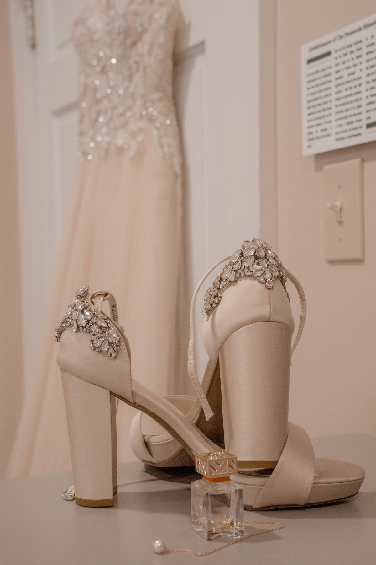 Crystal-heeled bridal sandals and a perfume bottle in front of the wedding dress hanging on the door — photo by Jimmy Warr
