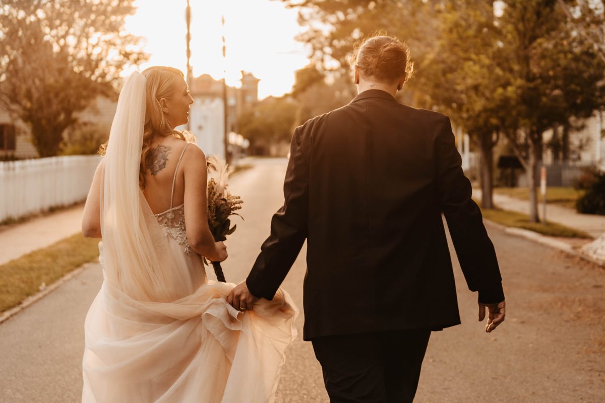 The couple walk away down a tree-lined street at sunset, the groom carrying her train — photo by Jimmy Warr