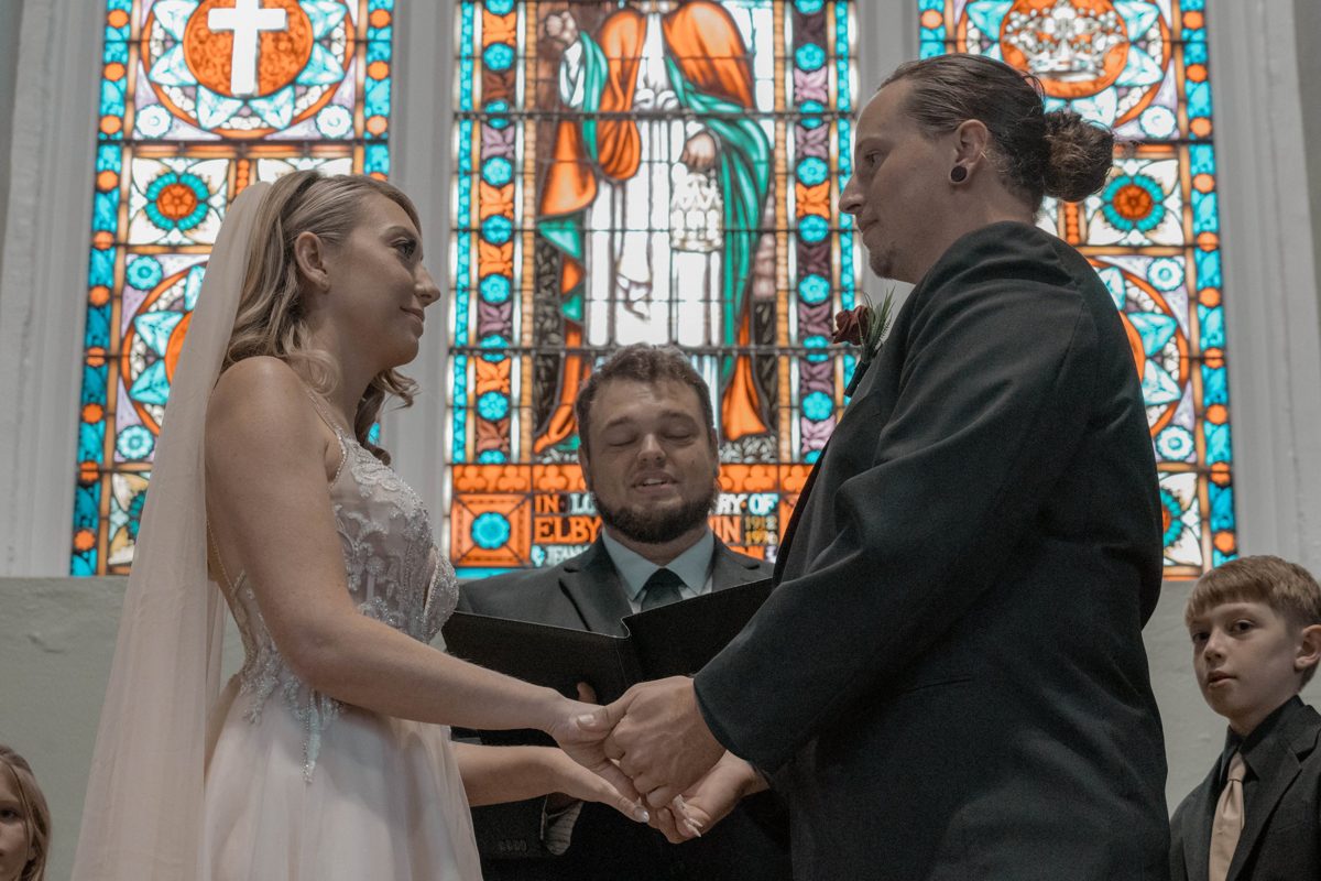 The couple hold hands during their vows beneath a stained-glass window, the officiant smiling between them — photo by Jimmy Warr