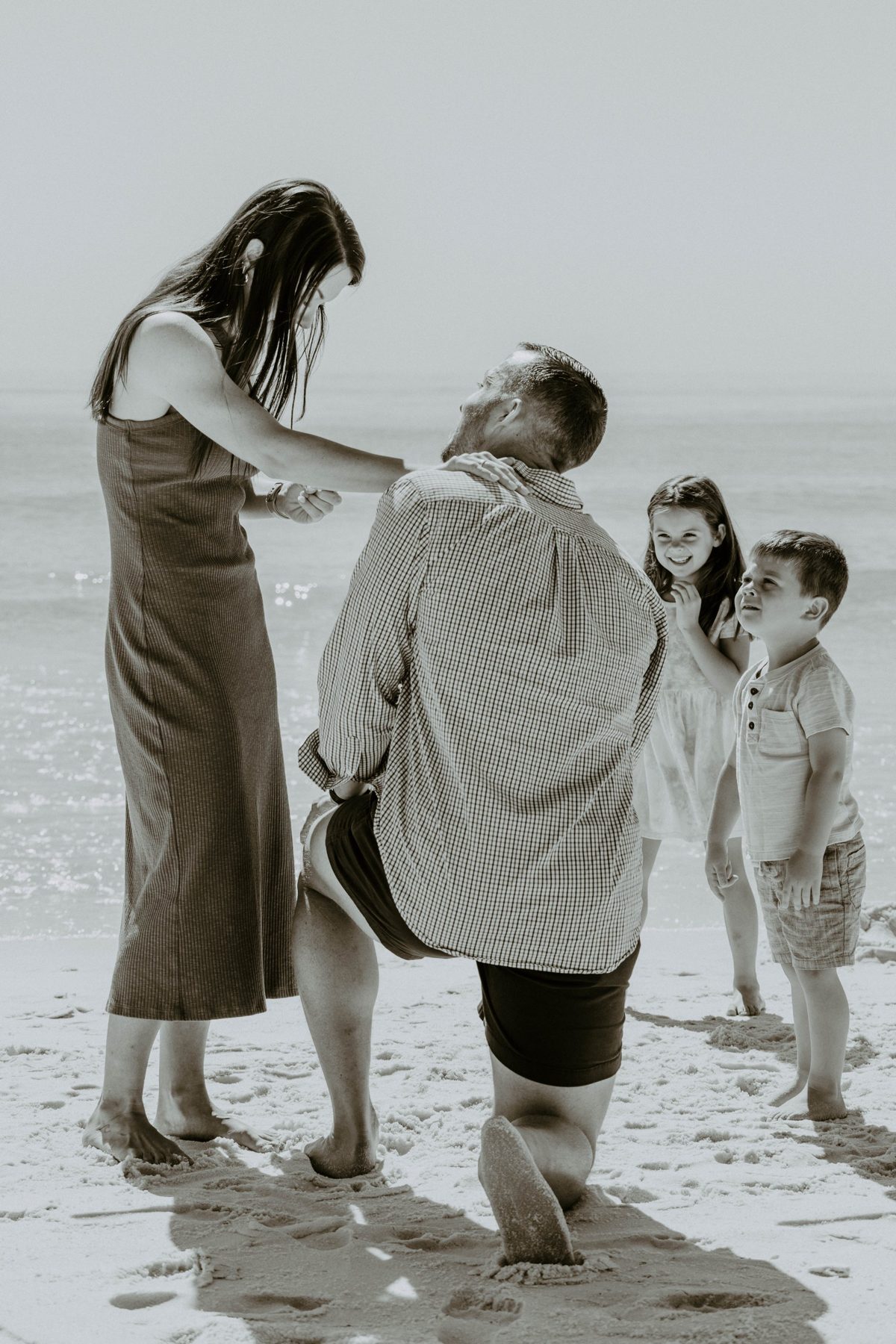 Seen from behind, he kneels as she looks at the ring, two children beaming up at them — photo by Jimmy Warr