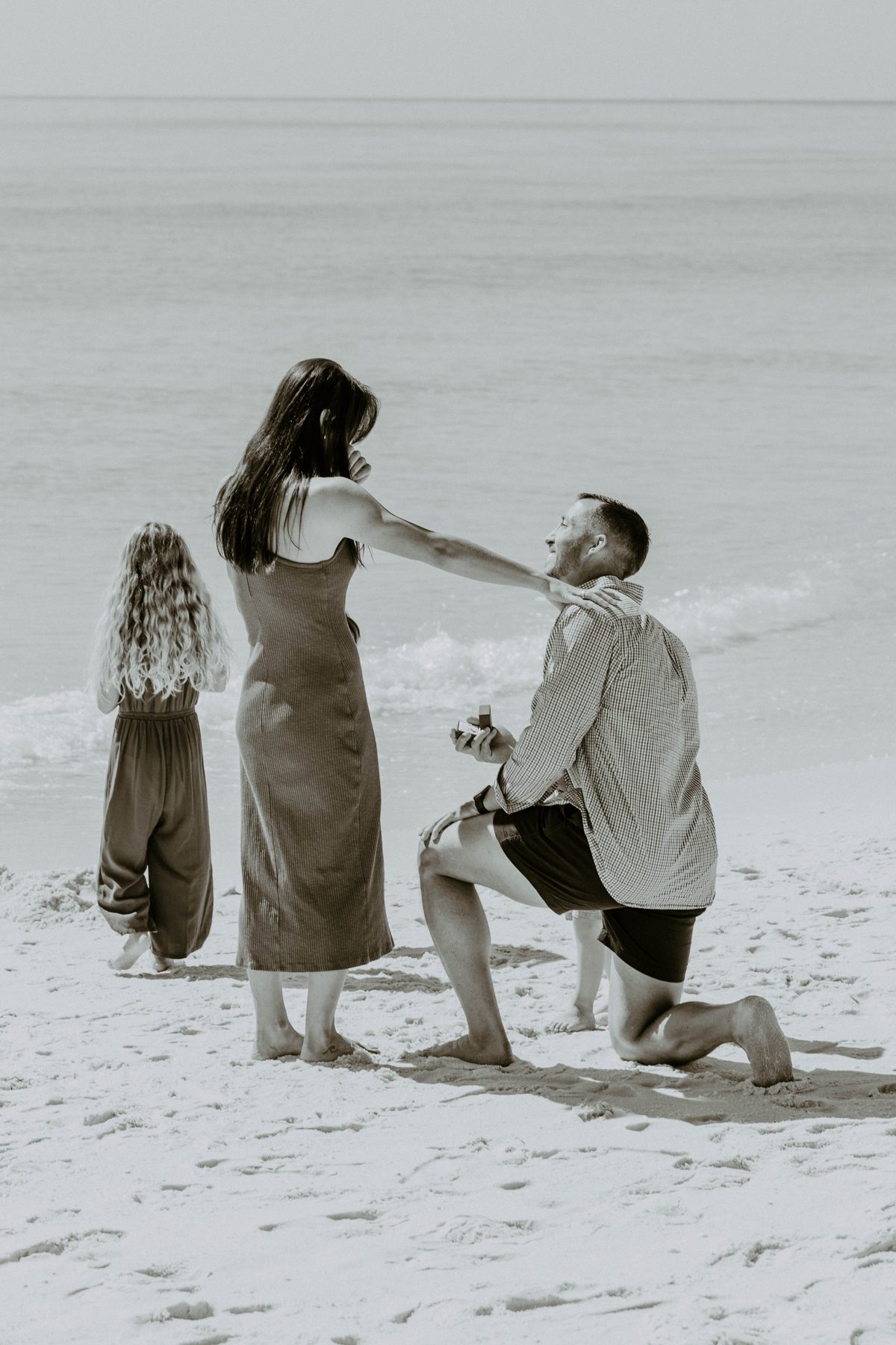 He kneels in the sand holding an open ring box while she reaches for his shoulder, the surf behind them — photo by Jimmy Warr
