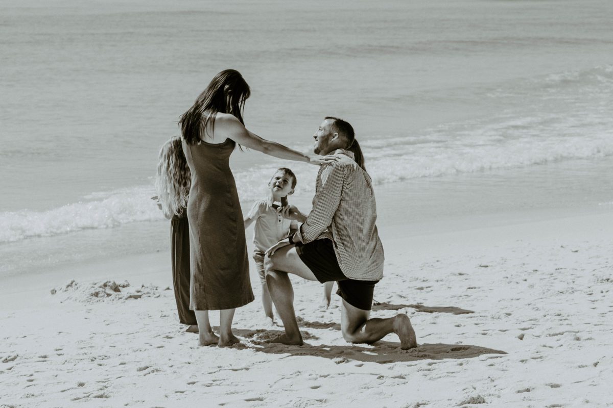 A man kneels on Pensacola Beach to propose as his partner rests a hand on his shoulder, the family's children close by at the water's edge — photo by Jimmy Warr