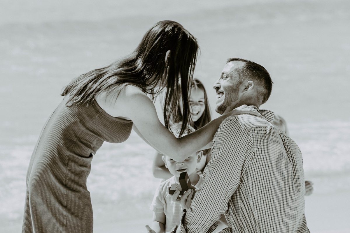 Black-and-white close moment: she leans in laughing as he kneels with the ring box, the children crowding in to watch — photo by Jimmy Warr