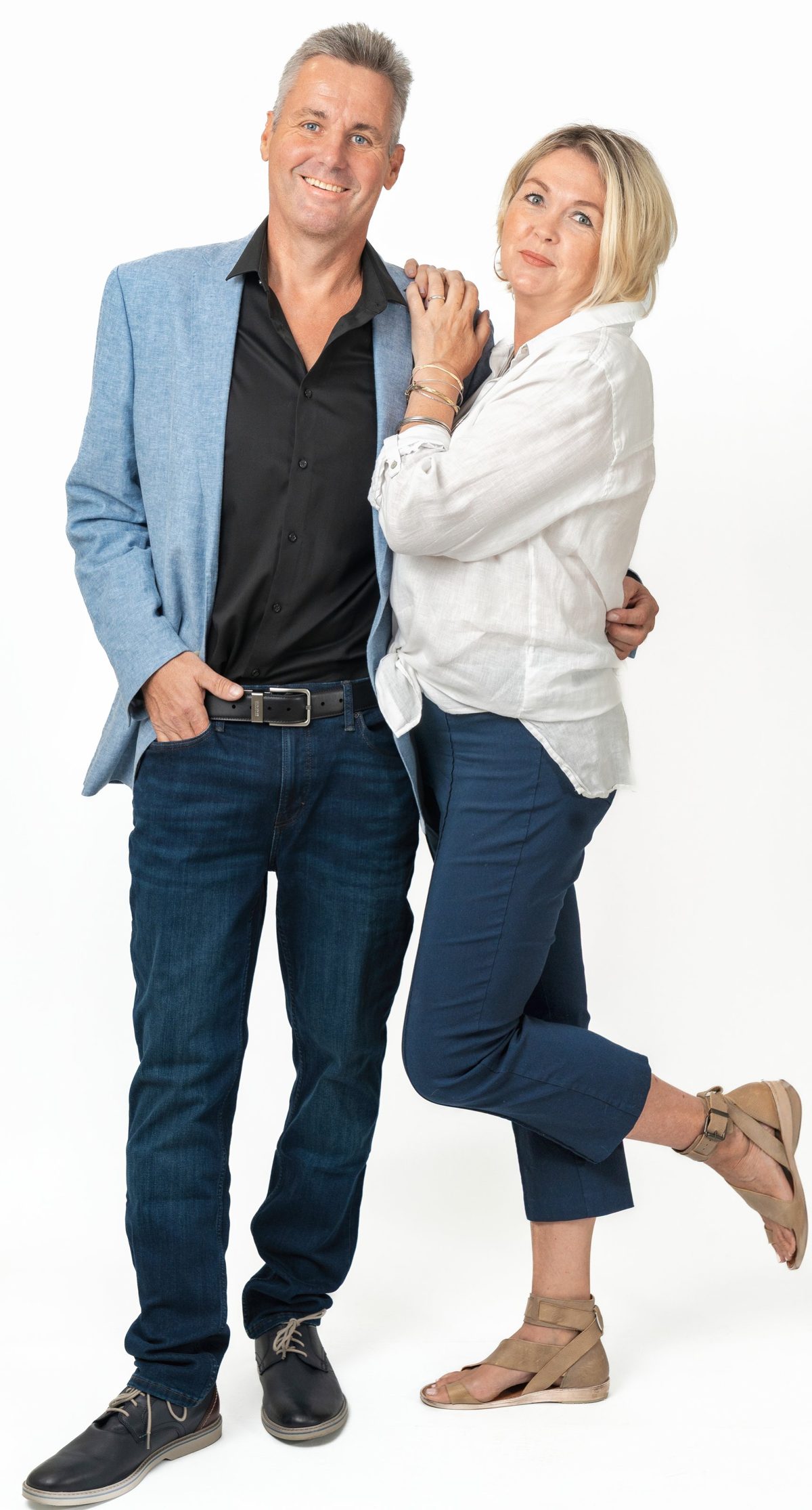Two realtors pose together on a white backdrop, he in a light blue blazer and black shirt, she in a white linen shirt with one foot kicked up behind her — photo by Jimmy Warr