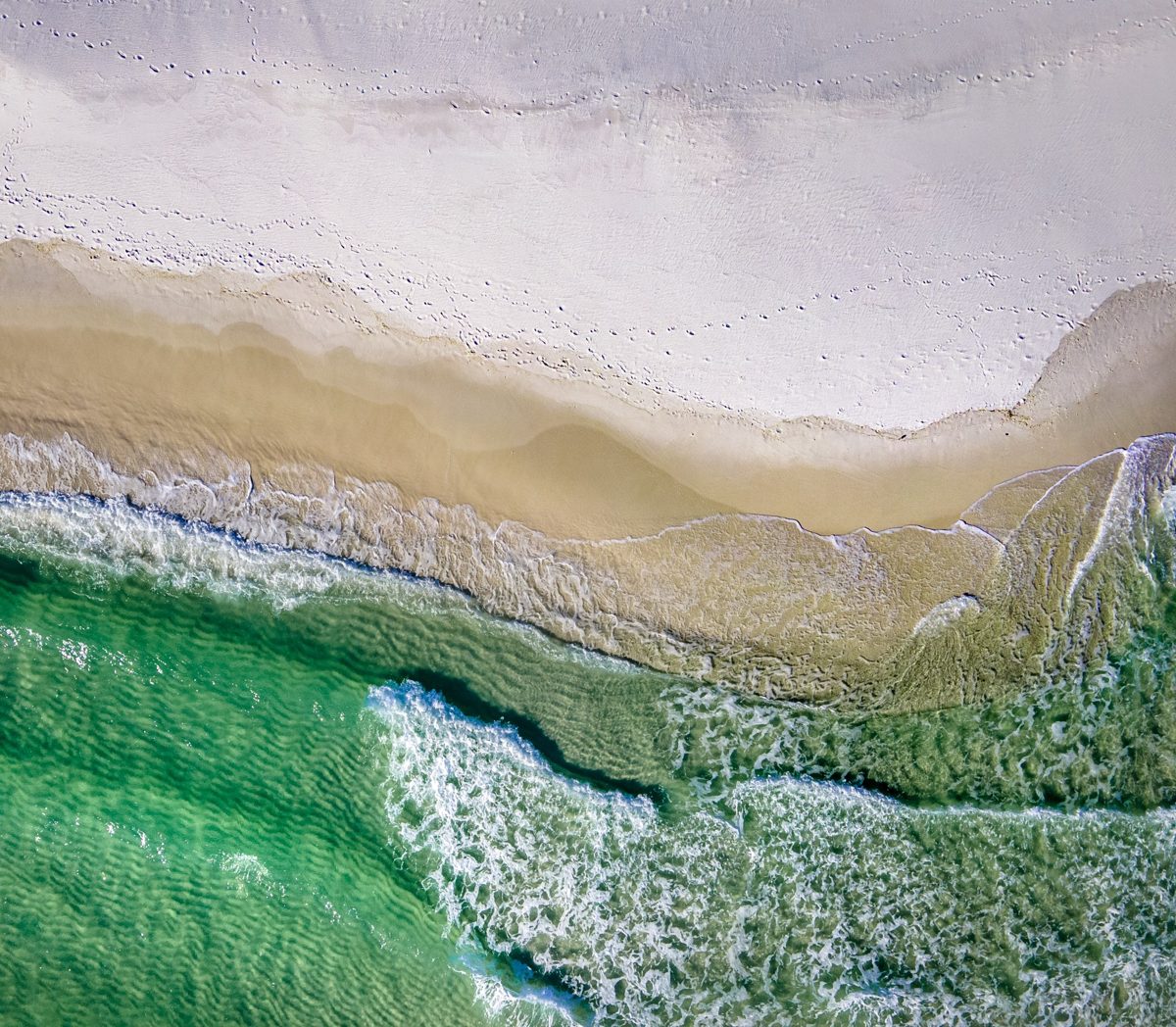 Straight-down aerial of the tide rolling onto Pensacola Beach: emerald water, white foam, and footprints in the sugar-white sand — photo by Jimmy Warr