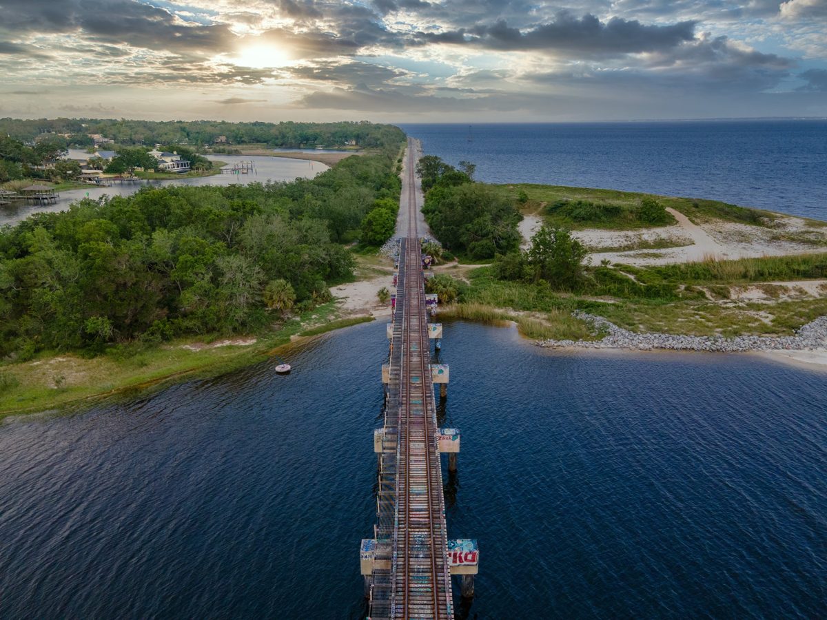 Aerial of a graffiti-covered railroad trestle running across the water toward the bay, the sunrise breaking through clouds over Pensacola — photo by Jimmy Warr