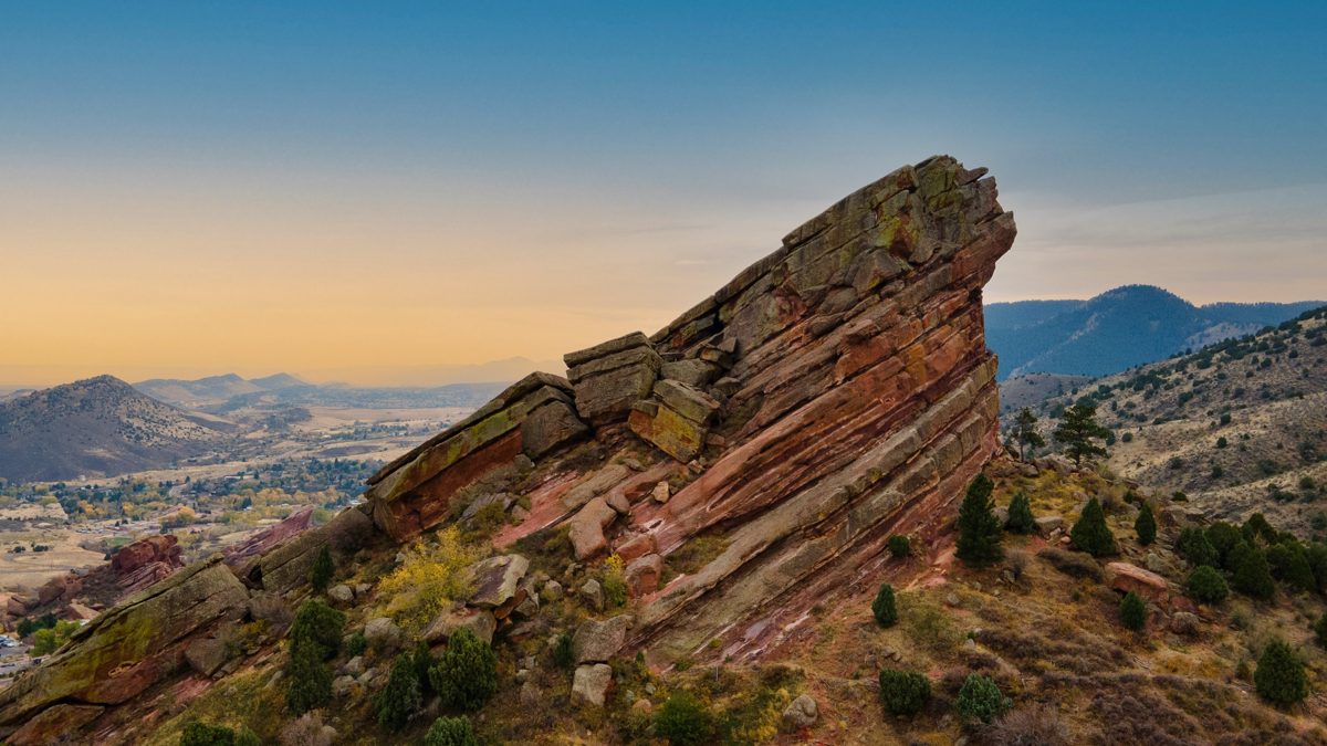 A tilted red sandstone fin at Red Rocks glows at sunrise, the foothills and Denver plain fading into gold haze — photo by Jimmy Warr