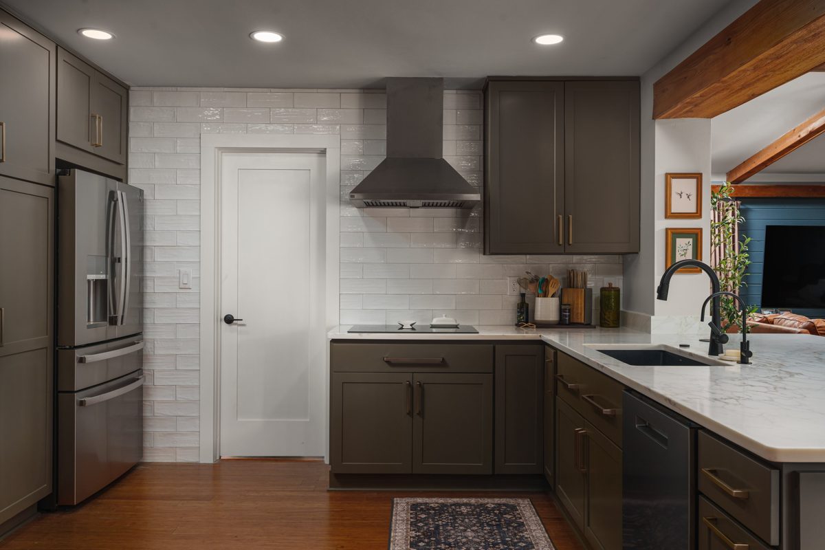 Kitchen wall of olive Shaker cabinets with brass pulls, a stainless chimney hood, and handmade white tile running floor to ceiling — photo by Jimmy Warr