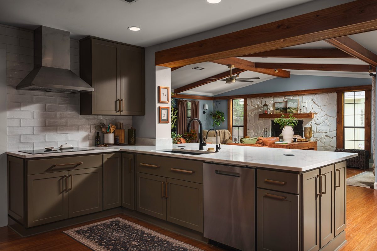 Olive Shaker cabinetry wrapping a marble-look peninsula, open beneath a cedar beam to the living room and white stone fireplace beyond — photo by Jimmy Warr