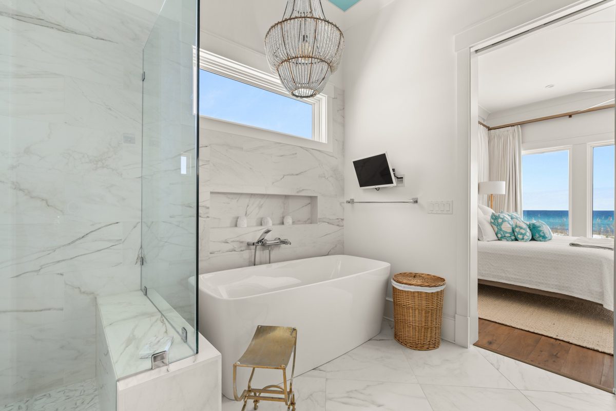 Marble bath with a freestanding tub under a beaded chandelier, the bedroom and Gulf visible through the doorway — photo by Jimmy Warr