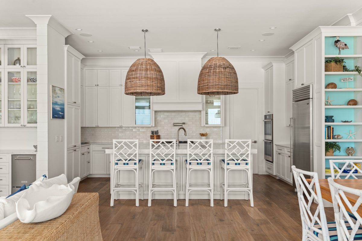 White Shaker kitchen under two oversized woven pendants, white bamboo-style stools, and a turquoise-backed bookcase — photo by Jimmy Warr