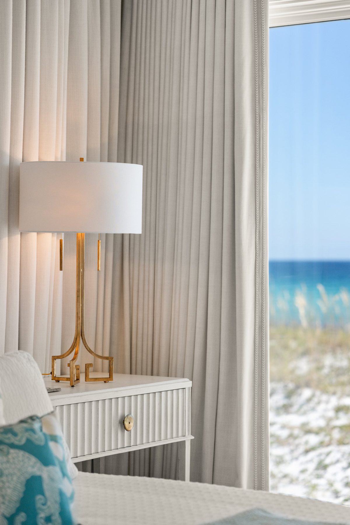 A gold fretwork lamp glowing on a fluted white nightstand, the Gulf soft through the window beside it — photo by Jimmy Warr