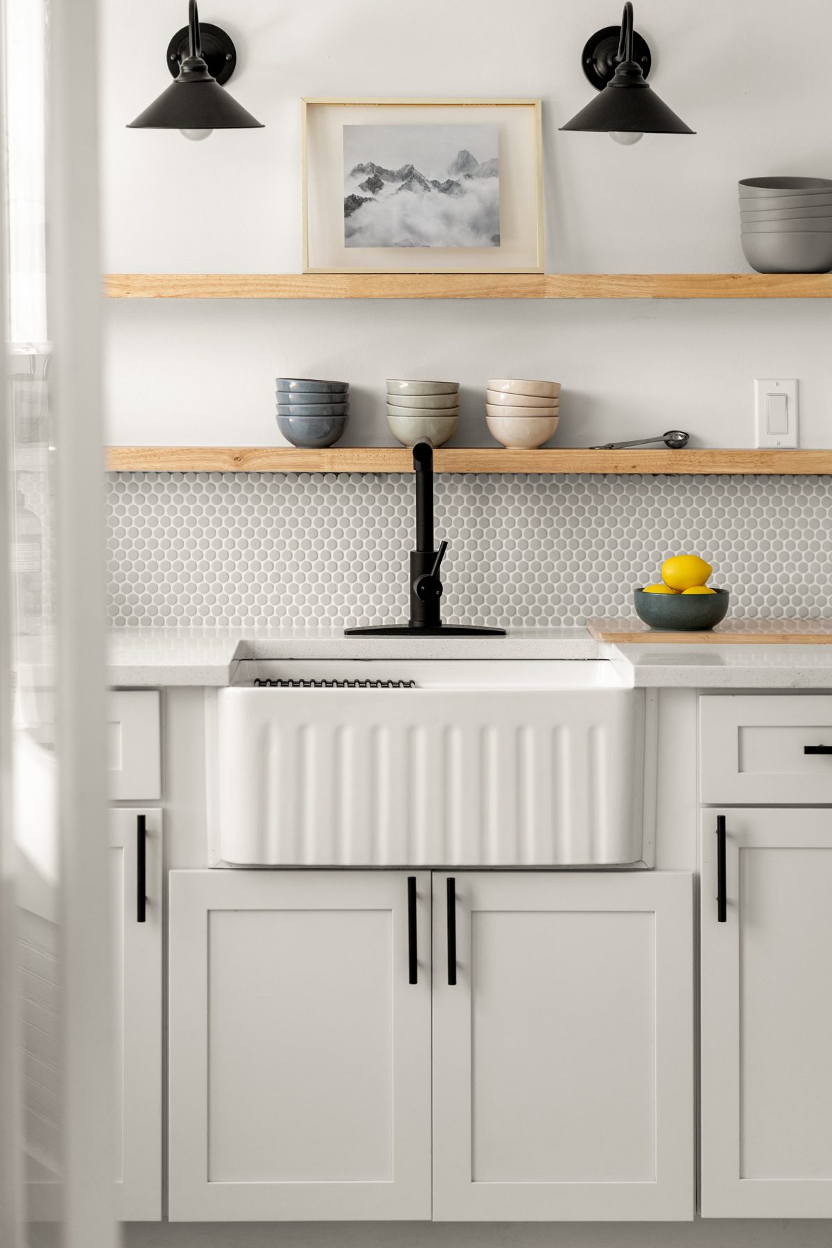 Fluted white farmhouse sink under open wood shelves, penny tile, and black barn sconces — photo by Jimmy Warr
