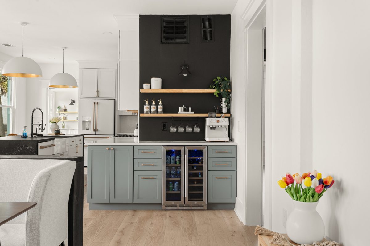 Coffee bar on a black accent wall with floating wood shelves over sage cabinets and a beverage fridge — photo by Jimmy Warr