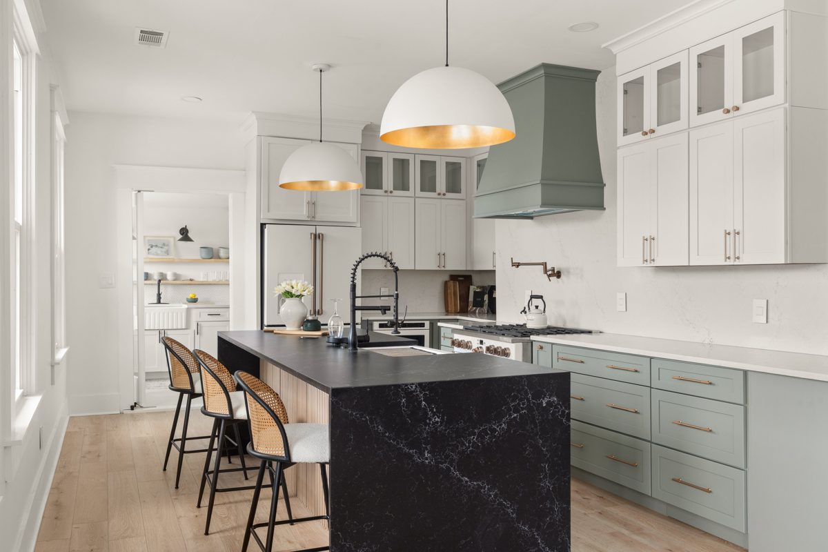 Kitchen with a black waterfall island, cane counter stools, and white-and-gold dome pendants beneath a sage range hood — photo by Jimmy Warr